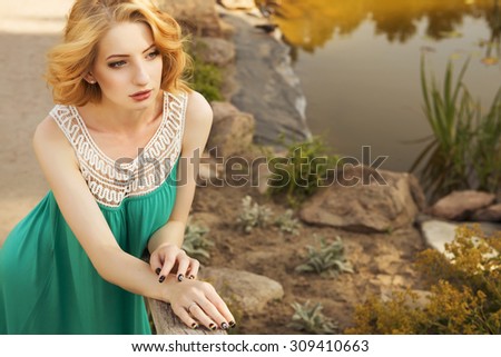Beautiful blonde woman with curly short bob hairstyle, delicate make up and red lips in green short dress at the park. Fashion sensual posing on summer sunset. Outdoors. Copy space