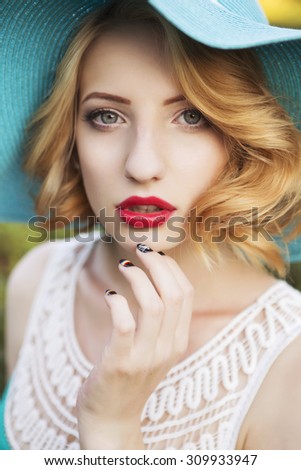 Beautiful blonde woman with curly short bob hairstyle, delicate make up and red lips in green short dress and a hat at the park. Fashion sensual posing on summer sunset. Outdoors