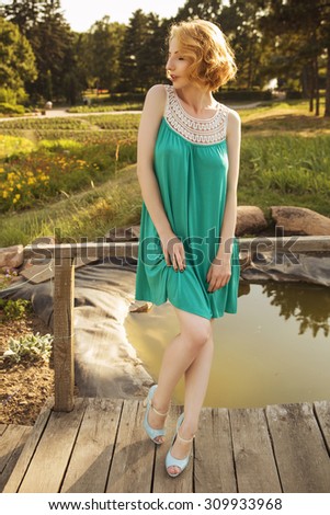 Beautiful blonde woman with curly short bob hairstyle, delicate make up and red lips in green short dress at the park. Fashion sensual posing on summer sunset. Outdoors