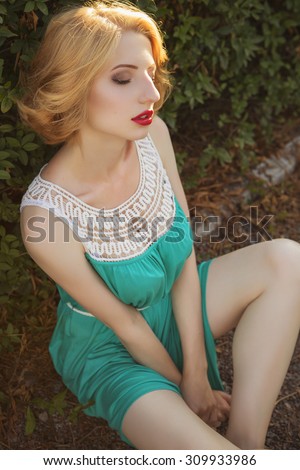 Beautiful blonde woman with curly short bob hairstyle, delicate make up and red lips in green short dress at the park. Fashion sensual posing on summer sunset. Outdoors