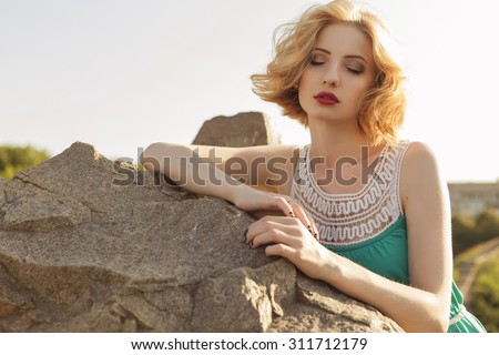 Beautiful blonde woman with curly short bob hairstyle, delicate make up and red lips in green short dress at the park. Fashion sensual posing on summer sunset. Outdoors. Copy space