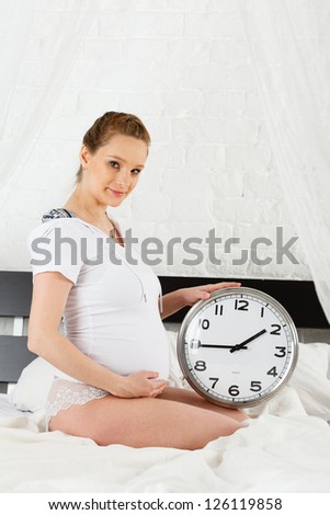 Pregnant woman with a clock sits on the house bed.