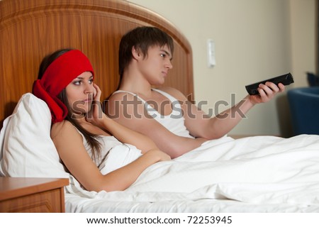 The unhappy young woman with a headache and her joyful husband  watching TV lie in bed. The family conflict.