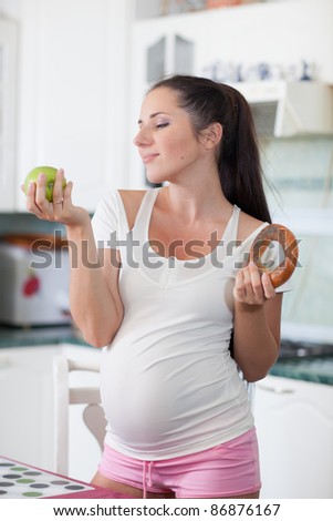 Young pregnant woman with apple and bagel in her hands in the house kitchen. Concept of healthy food.