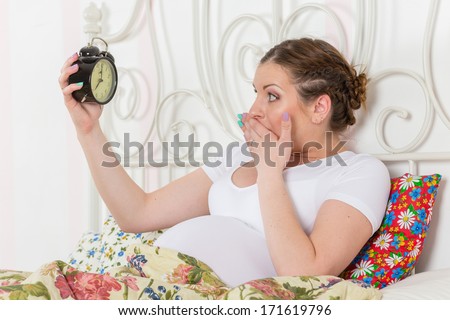Unhappy young pregnant  woman with horror looks at an alarm clock, lying in a house bed. Has overslept.