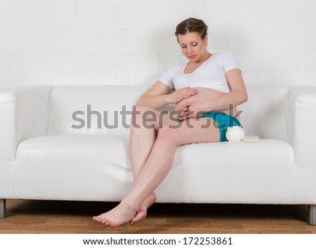 Young pregnant woman with cosmetic cream sits on a sofa in the room. Concept of body care.