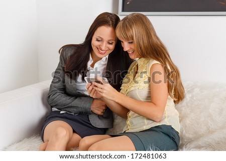 Two young business women with mobile phone sit on sofa in the office.