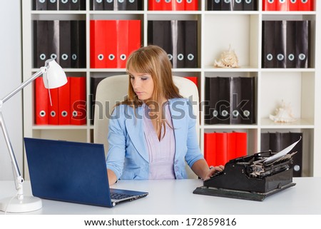 Young business woman  with notebook and old typewriter sits on workplace in the office. Progressing of technology.