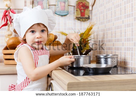 Little girl in apron and cap of the cook stands in the kitchen near cooker in the house. Mother\'s helper. 2 year old.