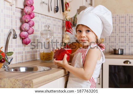 Little girl in apron and cap of the cook with an apple stands in the kitchen near sink at home. Mother\'s helper. 2 year old.