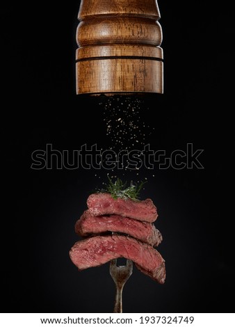 Roasted meat. Milled spices falling from pepper mill on grilled pieces of beef steak medium rare on fork on black background. Steak menu. Grilled menu.