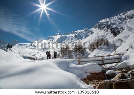 Tranquil early spring scene in the mountains with small group of people standing by snow covered bridge and flowing stream under magnificent snow covered mountain peaks and glaciers of the Alps.