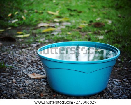 light blue color plastic water bucket on garden floor outdoor surrounding with green area environment and blue sky reflections on the water surface in the bucket