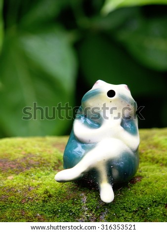 small green and white ceramic frog figure for garden decoration sitting on a brick cover with wet green moss next to garden pond with water plant background