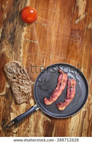 Fried Belly Bacon Rashers in Teflon Frying Pan with Bread Slice and Tomato alongside, on old, cracked, scratched, peeled off, obsolete Wooden Table surface.