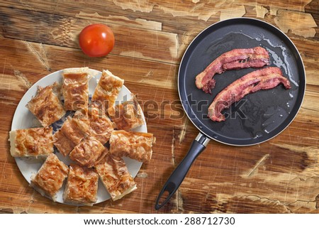 Fried Belly Bacon Rashers in Teflon Frying Pan with plateful of Serbian Gibanica cheese pie alongside, on old, cracked, scratched, peeled off, obsolete Wooden Table surface.