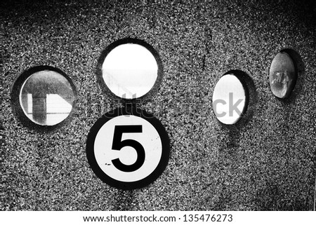 Black and white image of four round windows in a pebble dashed wall. Below the windows is a round sign of the number five