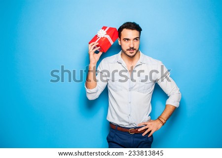 handsome man thinking on something, scratching his head with a christmas gift, on blue background