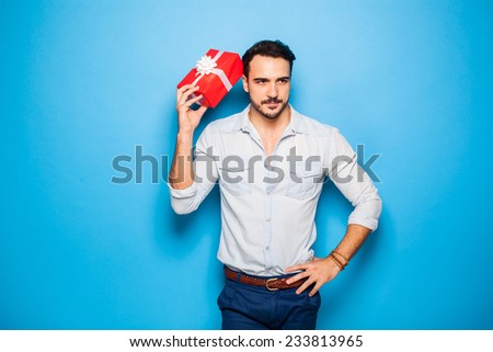 handsome man thinking on something, scratching his head with a christmas gift, on blue background