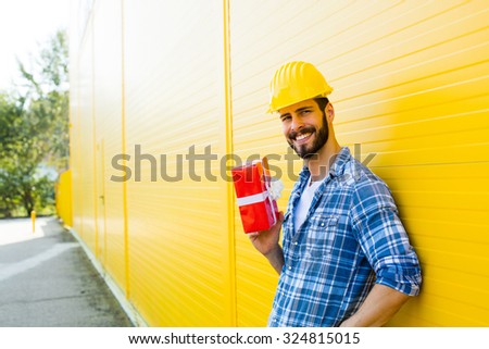 adult worker with yellow helmet and plaid shirt next to a wall smiling with a gift in hands