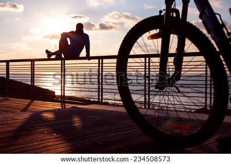 silhouette of young and active sportsman and his mountain bike sitting on the railing near the ocean and looking far away at the sunset