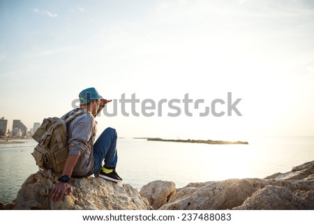 traveler with backpack sitting on the rocks near the sea on the beach in the city and looking far away at the horizon