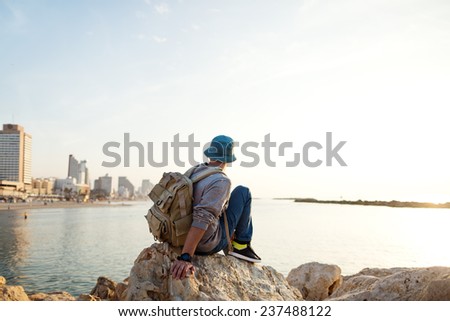 traveler with backpack sitting on the rocks near the sea on the beach in the city and looking far away