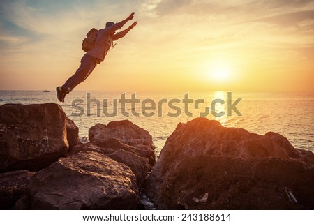 young brave man with backpack jumping over the rocks near the sea in sunset