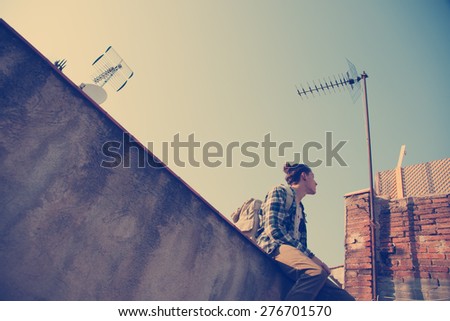 Young and brave man with backpack sitting on the edge of high roof and looking at antenna (intentional vintage color)