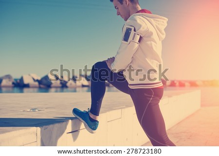 Handsome athlete doing legs stretching exercise after workout outdoors near the sea (intentional sun glare and vintage color)