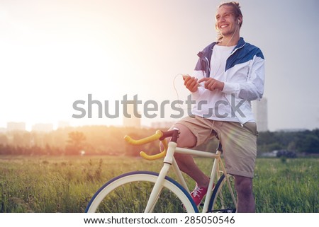 young smiling long hair man with bicycle in green field with smartphone (intentional sun glare and vintage colors, focus on phone)