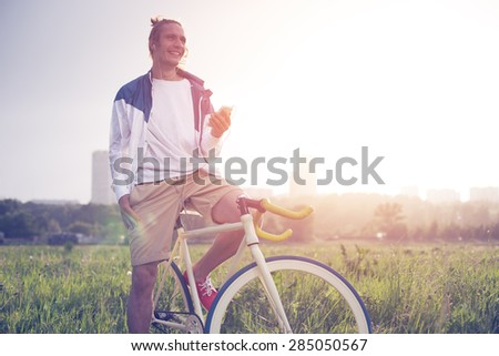 young smiling man with bicycle in green field with smartphone and earphones (intentional sun glare and vintage colors, focus on phone)