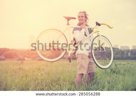Long hair man holding bicycle in green field at sunset (intentional sun glare and vintage color)