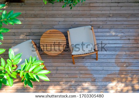 Top view of two stylish wooden chairs with cushions and a small wooden table at a balcony of a room in a resort, surrounded by garden 