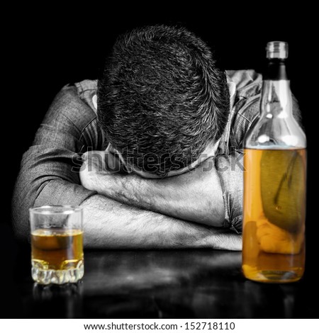 Black and white image of a drunk man sleeping with his head on a table and a bottle of whisky  (the bottle and glass have color)