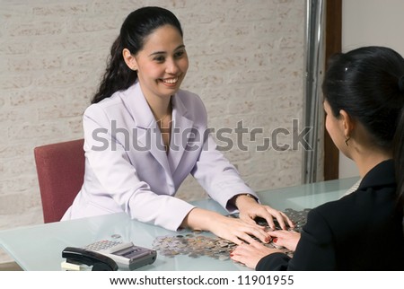 A shot of two young women sitting at glass desk covered in coins.  Their hands are on top of the desk moving the coins about.