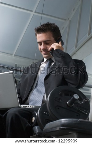 Man in a business suit sitting on a weight bench in a gym with his laptop and cell phone