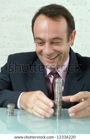Latin american businessman smiling broadly as he reaches for the larger of two stacks of coins in front of him on his desk.