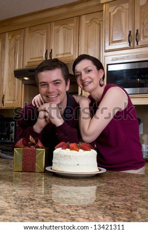 Attractive couple leaning on their kitchen counter which is covered with a cake and a present. They have a huge smile on their faces and are looking at the camera. Vertically framed shot.