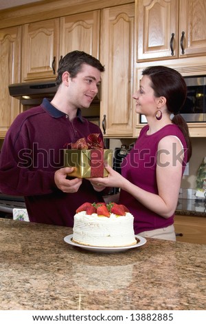Happy couple in a kitchen smiling near a cake. Both are holding a present while smiling at each . Vertically framed photograph.