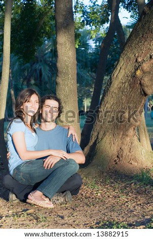 Happy, attractive couple sit in the woods. She sits on his lap a they both smile. Vertically framed photograph