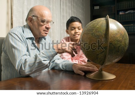 Grandfather and grandson are sitting at the table with a globe in front of them. Grandfather points to a location on the globe while grandson smiles beside him. This is a horizontally framed photo.