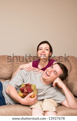 A smiling man holding a gift box lying on a smiling woman\'s lap on sofa.  Vertically framed shot.