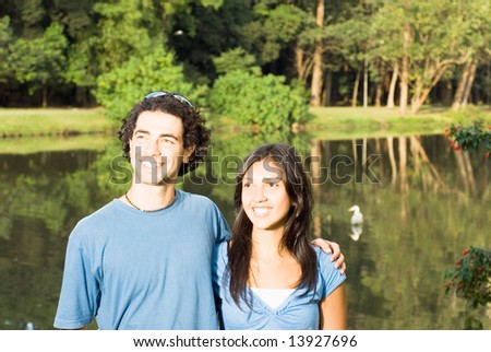Happy couple smiling in front of a pond. He has his arm around her as they stare off in the distance. Horizontally framed photograph
