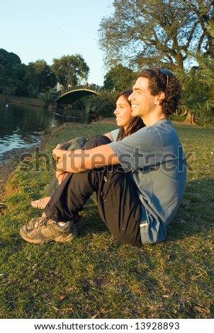 Couple sitting and talking by a pond. They are staring ahead and smiling. There is a bridge in the background. Vertically framed photograph