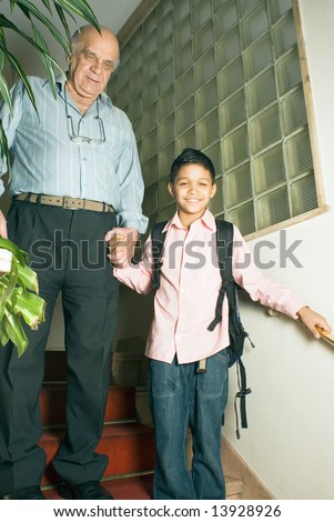 Grandfather and grandson are standing in the stairwell holding hands. Grandson is smiling with his backpack on as grandfather holds his hand. This is a vertically framed photo.