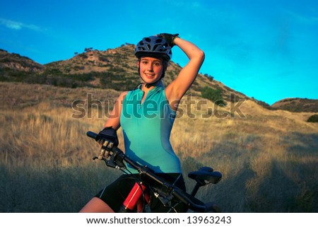 Attractive smiling female mountain-biker, wearing full cycle gear, straddling a mountain-bike adjusting her helmet.  Horizontally framed shot on with golden grasses and a blue sky in the background.