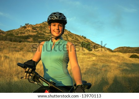 Attractive smiling female mountain-biker, wearing full cycle gear, straddling a mountain-bike looking off into the distance.  Horizontally framed shot.