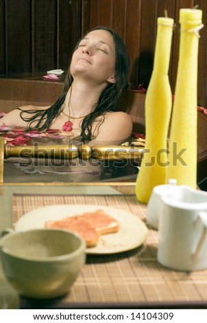 Woman relaxes in a spa tub filled with rose petals. She is closing her eyes and looks happy and relaxed. There are candles and lunch in the foreground. Vertically framed photograph