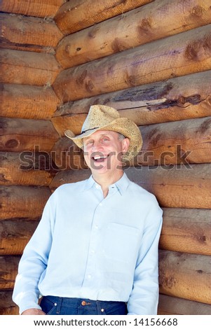 Laughing man in cowboy hat leaning against a log cabin. Vertically framed photograph